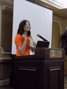 Woman in orange dress speaking at podium.